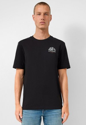 Junger Mann mit kurzen Haaren trägt ein schwarzes T-Shirt mit dem "Raw Beats Quitesense"-Logo auf der Brust, kombiniert mit hellblauen Jeans.