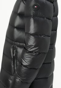 Zwarte gewatteerde pufferjack mouw met ritszak en rond Tommy Hilfiger logo patch met rood, wit en blauw vlagdetail.