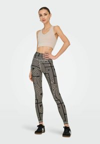 Leggings negros con patrones geométricos blancos, combinados con un top corto beige. La modelo está de pie con las manos en las caderas, vistiendo zapatillas negras.