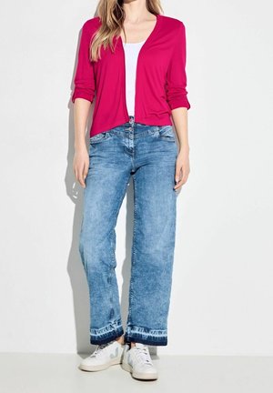 Vrouw met een felroze open cardigan, wit topje, wijd uitlopende blauwe spijkerbroek met donkere zomen en witte sneakers tegen een effen achtergrond.