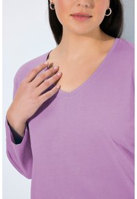 Halenka s dlouhým rukávem ve tvaru V ze světle levandulové bavlny s jemnou texturou. Ruka modelky spočívá poblíž výstřihu a ukazuje přirozený lak na nehty.