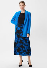 Blaues Blazer über einem schwarzen Oberteil, kombiniert mit einem fließenden schwarzen Rock mit einem blauen abstrakten Muster. Schwarze High Heels ergänzen das Outfit.