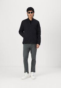 Black zip-up jacket with a stand collar, paired with gray slim-fit pants and white sneakers, standing against a plain background.