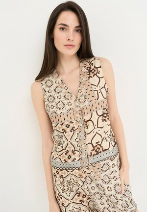 Ung kvinde med langt mørkt hår iført en ærmeløs beige og brun mønstret kjole, stående foran en ensartet hvid baggrund.