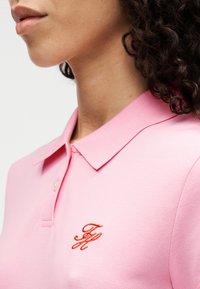 Ροζ πόλο πουκάμισο με ριμπ γιακά και κοντά μανίκια. Διαθέτει περίπλοκο κεντητό λογότυπο σε πορτοκαλί στο αριστερό μέρος του στήθους.