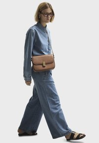 Brun läderväska med flap och guldfärgade detaljer, buren av en person i en blå jeansskjorta och vida jeans, som går.