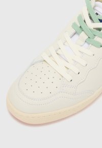 Cream-colored sneaker with a textured rubber sole, perforated toe, and light green laces. Features glossy stitching and a smooth leather upper.