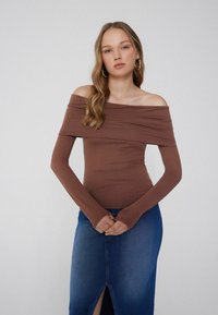 Junge Frau mit langen blonden Haaren, die ein braunes, schulterfreies Langarmoberteil und einen blauen Jeansrock mit vorderem Schlitz trägt, steht vor einem schlichten Hintergrund.