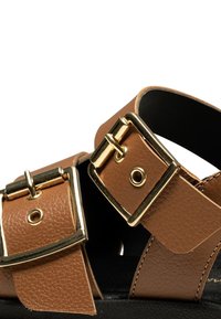 Gros plan sur des brides de sandales en cuir marron avec de grandes boucles rectangulaires en métal doré et un fond de semelle noir.