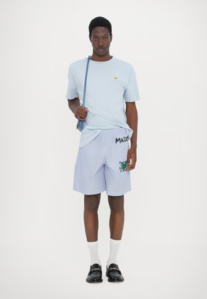 Jasnoniebieska bawełniana koszulka z małym żółtym logo, w zestawie z niebieskimi i białymi spodniami w paski z tekstem i haftowanymi grafikami. Czarne mokasyny.