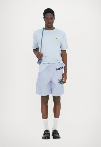 Helle blaue Baumwoll-T-Shirt mit kleinem gelbem Logo, kombiniert mit blau-weißen gestreiften Shorts mit Text und gestickten Grafiken. Schwarze Slipper.