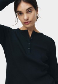 Sort ribbet langærmet top med henley-udskæring, der har en knaplukning og blød tekstur, båret med en arm hævet.