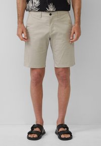 Shorts beige en coton avec devant plat, poches latérales et fermeture à bouton, associés à des sandales noires à doubles lanières.