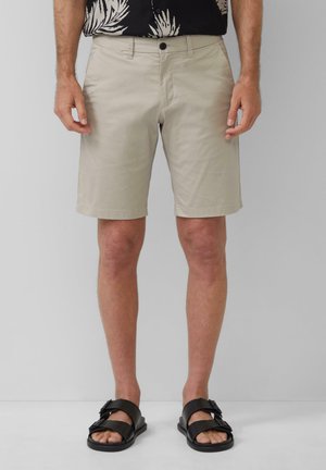 Beige Shorts aus Baumwolle mit flachem Bund, seitlichen Taschen und einem Knopfverschluss, kombiniert mit schwarzen Sandalen mit doppelten Riemen.