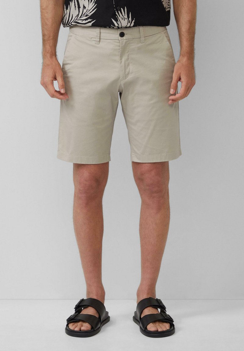 Shorts beige en coton avec devant plat, poches latérales et fermeture à bouton, associés à des sandales noires à doubles lanières.
