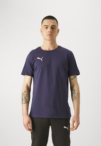 Mørkeblå bomull t-skjorte med rund hals og korte ermer. Har en hvit Puma-logo på venstre bryst. Modellen har på seg svarte bukser med detaljer.