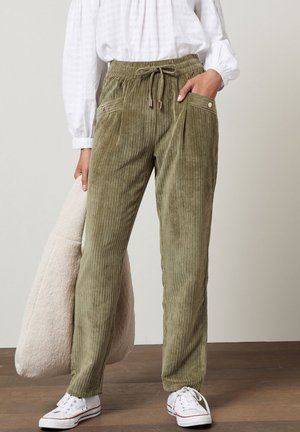 Pantalon en velours côtelé vert olive avec taille élastique à cordon, poches avant, porté avec des baskets blanches et un chemisier blanc, tenant un sac beige texturé.