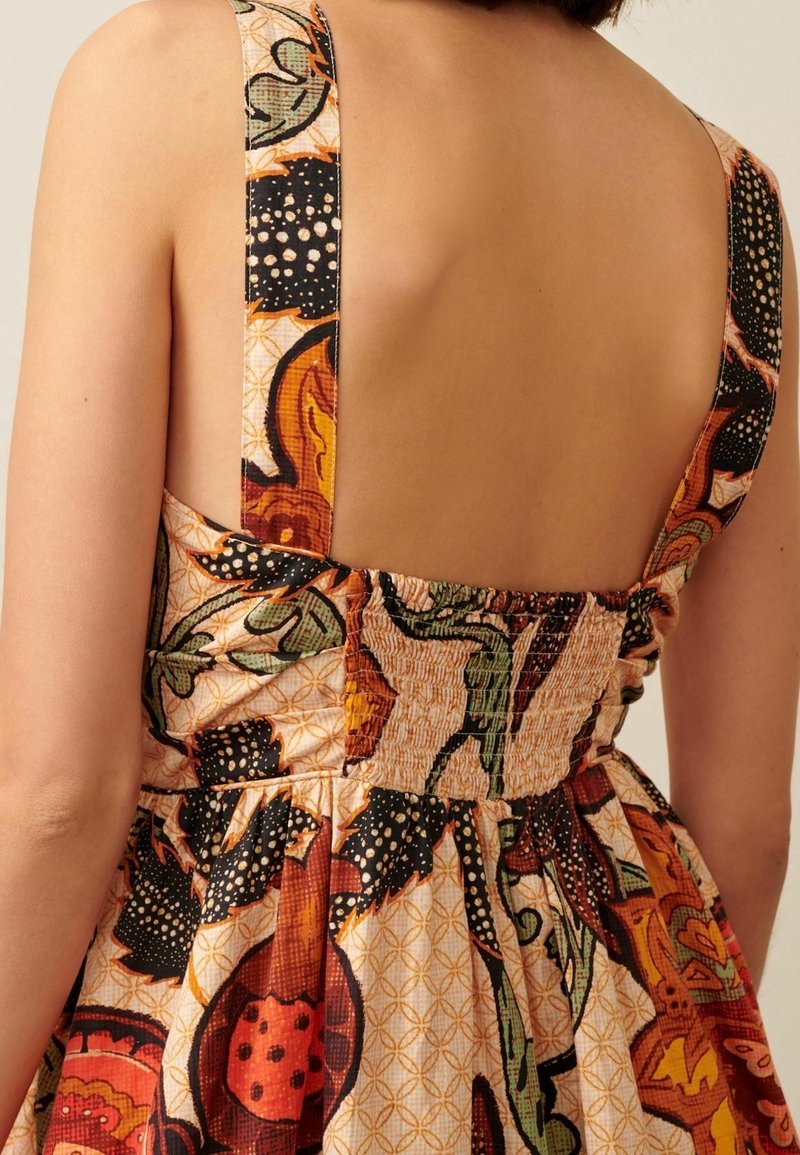 Femme portant une robe sans manches avec de larges bretelles et un motif floral coloré, montrant un dos nu et un tissu froncé à la taille.