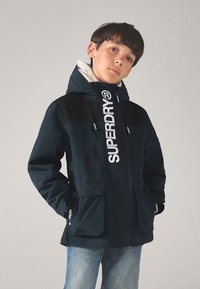 Tumesinine jakk valge logoga, mustade detailide, tõmblukkude ja kahe esitaskuga. Kapuutsiga disain pehme, tekstureeritud sisuga.