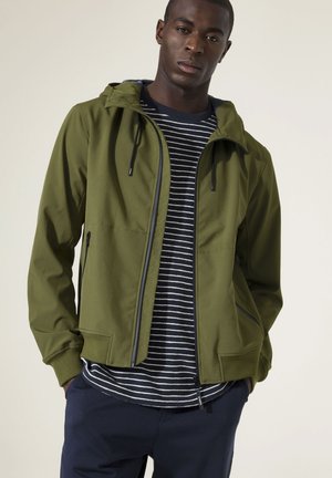 Man draagt een olijfgroene capuchonjas over een zwart-wit gestreept shirt en een donkere broek, staat met zijn handen in de zakken tegen een effen achtergrond.