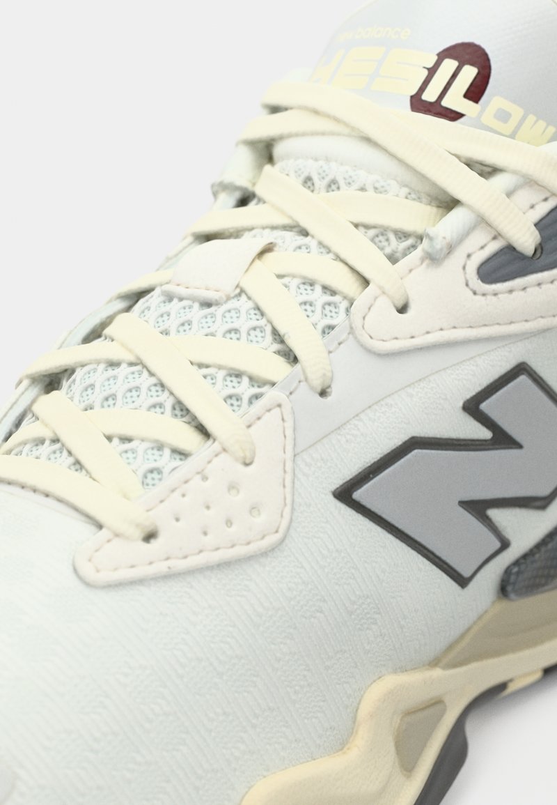 Hellgrauer Sportschuh mit strukturierter Oberseite, Netzpaneelen, elfenbeinfarbenen Schnürsenkeln und einem großen grauen "N"-Logo an der Seite.
