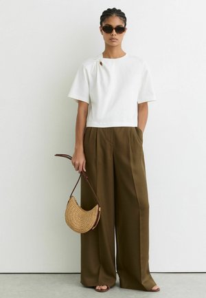 Femeie purtând un top alb cu mâneci scurte, pantaloni largi kaki, ochelari de soare închiși la culoare, ținând pe lângă ea o geantă împletită în formă semilunară.