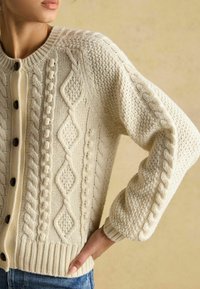 Svetlo béžový kardigán s károvaným pletením a textúrovanými vzormi, s veľkými hnedými gombíkmi a rebrovanými manžetami. Mäkko, hrubé pletené materiály.