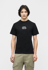 Νέος άνδρας με μεσαίου μήκους σκουρόχρωμα μαλλιά, φορώντας μαύρο μπλουζάκι με την επιγραφή "Replay Custom Service" και μπεζ cargo παντελόνια, στέκεται μπροστά σε απλό φόντο.