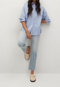 Light blue striped button-up shirt, rolled sleeves, paired with light denim jeans and beige slip-on shoes. White background.