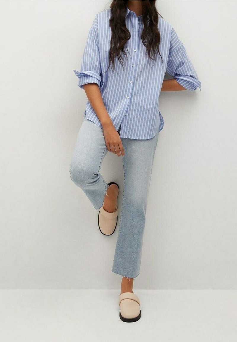 Light blue striped button-up shirt, rolled sleeves, paired with light denim jeans and beige slip-on shoes. White background.