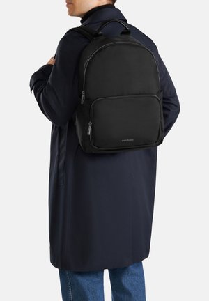 Person trägt einen marineblauen Mantel und blaue Jeans und trägt einen schwarzen Gino Rossi Rucksack mit vorderer Reißverschlusstasche auf dem Rücken.