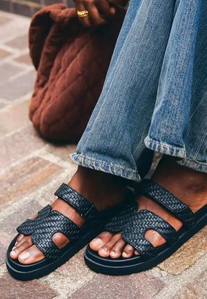 Sandales tissées noires portées avec un jean en denim bleu clair, à côté d'un sac matelassé marron sur un pavé en briques.