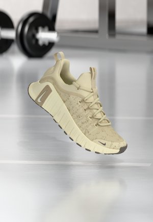 Chaussure de course Nike Free beige clair avec une semelle épaisse et des lacets, présentée dans une salle de sport avec du matériel de musculation flou en arrière-plan.