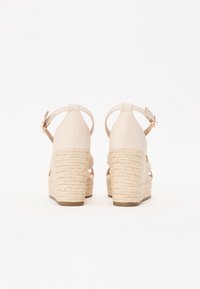 Beige kile-sandaler med en flettet jute-helle, ankelstropp og spenne-detail. Glatt lær-overdel med åpen design på hælen.