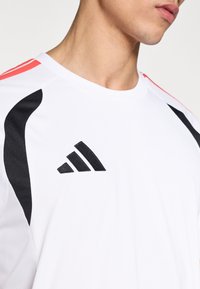 Λευκό αθλητικό πουκάμισο με μαύρες και κόκκινες λεπτομέρειες, με υφή δικτυωτού κ αι κεντημένο μαύρο λογότυπο. Στρογγυλή λαιμόκοψη και κοντά μανίκια.