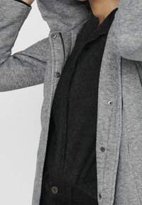 Un capo d'abbigliamento grigio e strutturato con cappuccio e chiusure a pressione, sovrapposto a un top nero e soffice. Visibili jeans scuri nella parte inferiore.