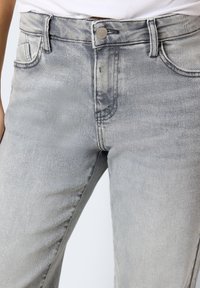 Gros plan sur un jean en denim gris clair porté avec un haut blanc côtelé rentré, montrant le bouton avant et les poches.