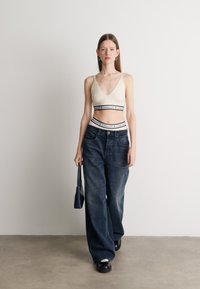 Svetlá béžová pruhovaná crop top s tenkými ramienkami, v kombinácii s voľnými modrými denimovými džínsami a malou čiernou kabelkou. Hladká textúra a pohodlný strih.