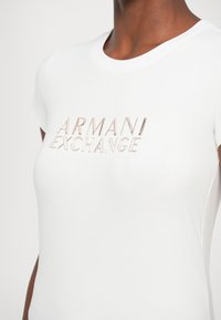 Biely bavlnený tričko s okrúhlym výstrihom a krátkymi rukávmi, s metalickým logom "ARMANI EXCHANGE" na prednej strane. Hladká textúra.
