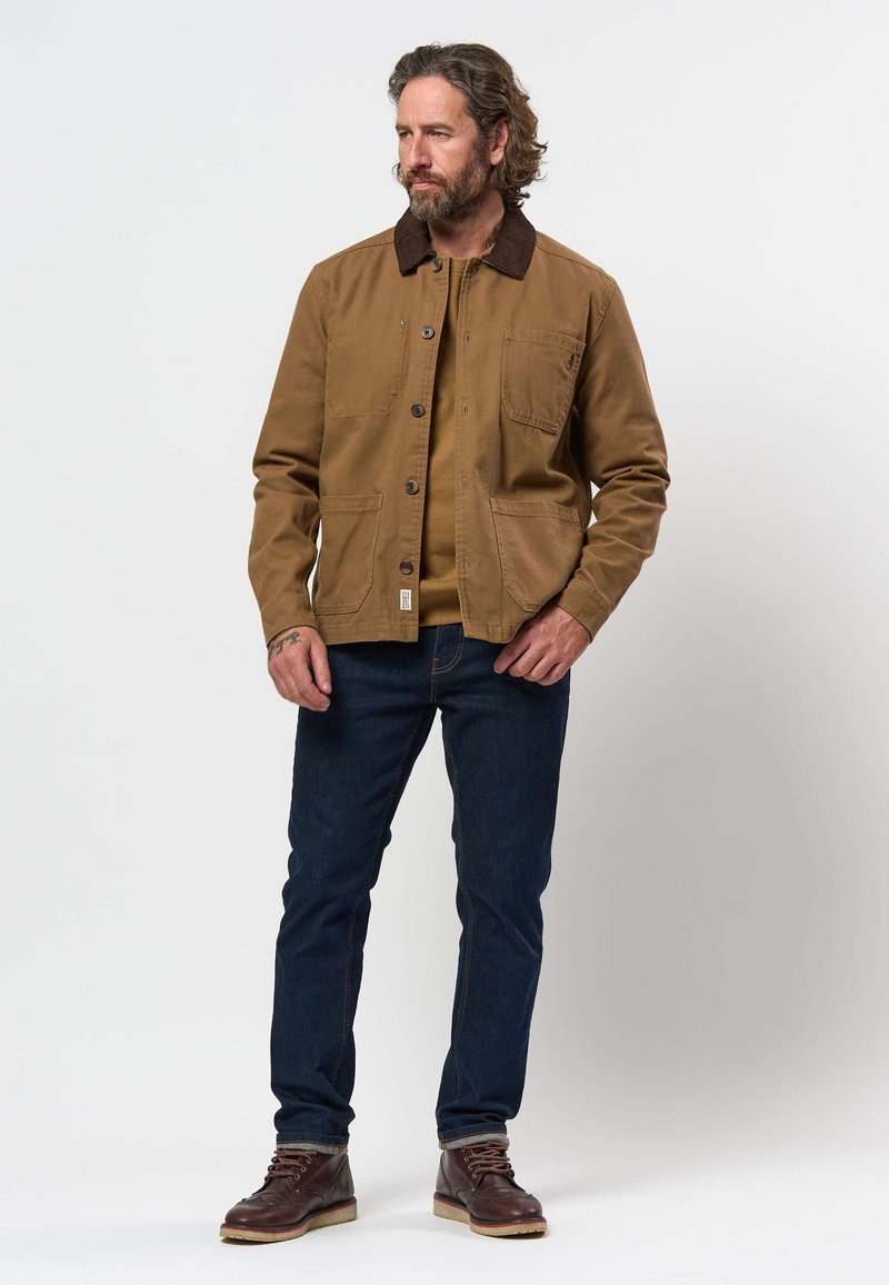 Homme avec une veste marron, un jean bleu et des bottes marron se tient devant un fond blanc en regardant sur le côté.