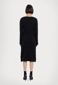 Femme aux cheveux courts et foncés, portant une robe midi noire à manches longues, des chaussettes noires et des chaussures noires à talons hauts, debout, dos tourné.