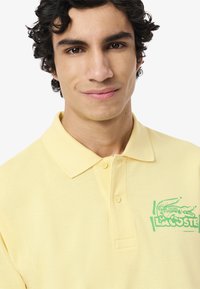 Polo giallo realizzato in tessuto testurizzato con colletto, dotato di logo Lacoste verde sul lato sinistro del petto e chiusura con due bottoni.