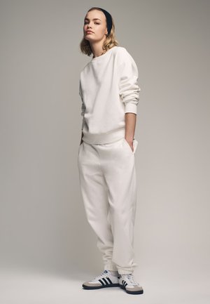 Junge Frau trägt weißen Sweatshirt und Jogginghose, schwarzes Stirnband und weiße Sneakers mit schwarzen Streifen, steht mit Händen in den Taschen.