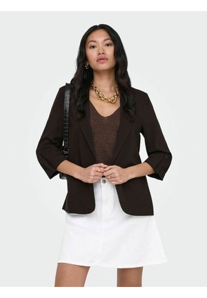 Femme portant un blazer marron foncé, un top marron, une jupe blanche, un collier chaîne doré et un sac à main noir, debout devant un fond uni.