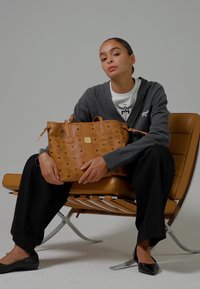 Donna in cardigan grigio e pantaloni neri è seduta su una sedia beige, mentre tiene una grande borsa a tracolla in pelle marrone con un logo decorato.