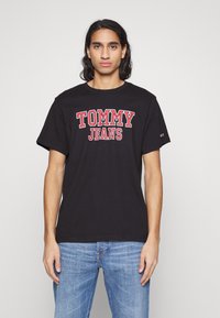 Juodas medvilninis marškinėliai su ryškiu raudonu užrašu "TOMMY JEANS" per krūtinę, trumpomis rankovėmis ir apvaliu kaklu.