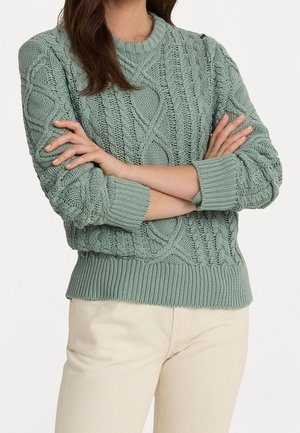 Pull en maille torsadée de couleur vert pastel, avec un col et des poignets côtelés, présentant des motifs texturés complexes et une silhouette ajustée.
