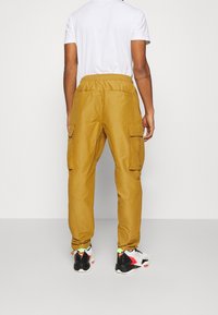 Žluté cargo kalhoty s elastickým páskem, dvěma bočními kapsami a hladkou texturou. Nošeny s bílým tričkem a vícebarevnými teniskami.