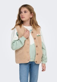 Beeži fleecevesti, millel on nupulukk, ümar kaelus, küljetaskud ja värvikas plaastritükk. Kantud helerohelise ja valge kampsuni peal.