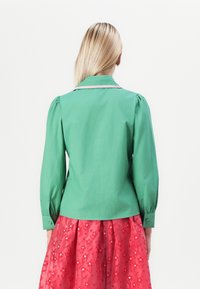 Camisa verde con cuello de pico y mangas abullonadas, con un acento rosa en el cuello; combinada con una falda roja con estampado floral.